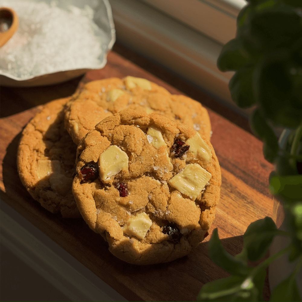 Cookies med hvit sjokolade og tranebær på trefjøl ved vindu, basilikum i forgrunnen