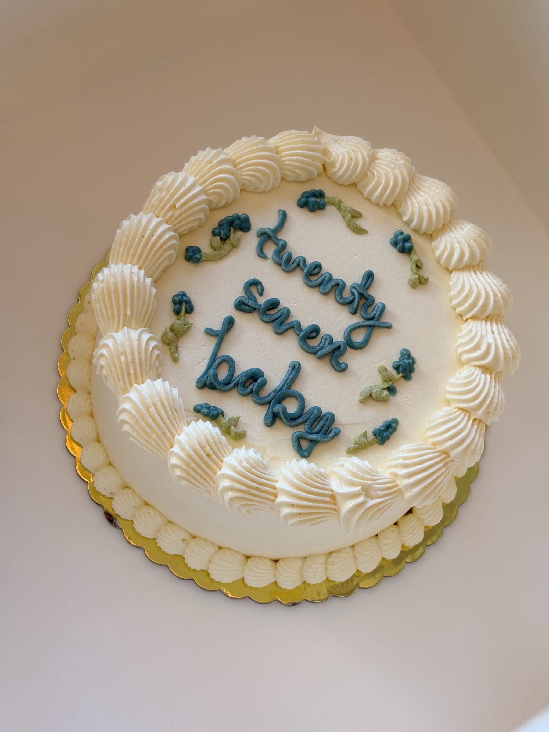 Vintage-stil bursdagskake i hvit smørkrem med skjellpiping rundt kanten og «twenty seven baby» skrevet i blågrønn krem